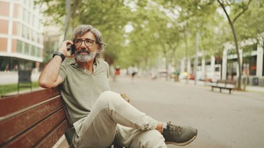 Gri saçlı ve sakallı, sıradan kıyafetler giyen orta yaşlı bir adam bankta oturuyor ve cep telefonu kullanıyor. Gözlüklü, akıllı telefondan konuşan olgun bir beyefendi.