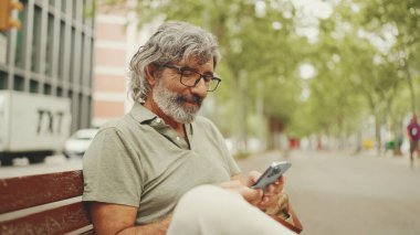 Dost canlısı, orta yaşlı, gri saçlı, sakallı sıradan kıyafetler giyen bir adam bankta oturuyor. Gözlüklü olgun bir beyefendi kameraya bakar ve gülümser.