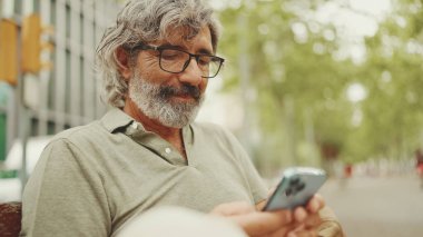 Dost canlısı, orta yaşlı, gri saçlı, sakallı sıradan kıyafetler giyen bir adam bankta oturuyor. Gözlüklü olgun bir beyefendi kameraya bakar ve gülümser.