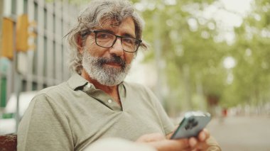Dost canlısı, orta yaşlı, gri saçlı, sakallı sıradan kıyafetler giyen bir adam bankta oturuyor. Gözlüklü olgun bir beyefendi kameraya bakar ve gülümser.