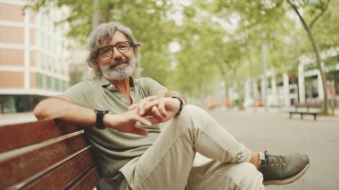 Gri saçlı, sakallı, sıradan kıyafetler giyen düşünceli orta yaşlı bir adam bankta oturuyor. Gözlüklü, olgun bir beyefendi başını çevirip kameraya bakıyor.