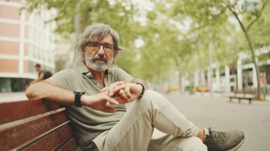 Gri saçlı, sakallı, sıradan kıyafetler giyen düşünceli orta yaşlı bir adam bankta oturuyor. Gözlüklü, olgun bir beyefendi başını çevirip kameraya bakıyor.