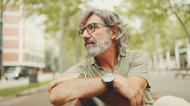 Gri saçlı, sakallı, sıradan kıyafetler giyen düşünceli orta yaşlı bir adam bankta oturuyor. Gözlüklü, olgun bir beyefendi başını çevirip kameraya bakıyor.