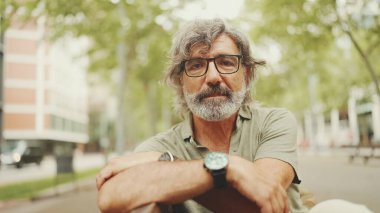 Gri saçlı, sakallı, sıradan kıyafetler giyen düşünceli orta yaşlı bir adam bankta oturuyor. Gözlüklü, olgun bir beyefendi başını çevirip kameraya bakıyor.