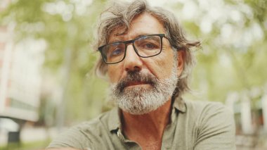 Gri saçlı, sakallı, sıradan kıyafetler giyen düşünceli orta yaşlı bir adam bankta oturuyor. Gözlüklü, olgun bir beyefendi başını çevirip kameraya bakıyor.