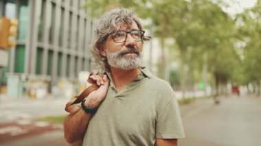 Gri saçlı, sakallı orta yaşlı bir adam yürüyor ve etrafına bakıyor. Omzunda çanta olan gözlüklü olgun bir beyefendi şehir manzarası arka planında meydanda yürüyor.