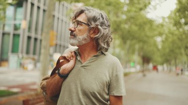 Gri saçlı, sakallı orta yaşlı bir adam yürüyor ve etrafına bakıyor. Omzunda çanta olan gözlüklü olgun bir beyefendi şehir manzarası arka planında meydanda yürüyor.