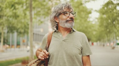 Gri saçlı, sakallı orta yaşlı bir adam yürüyor ve etrafına bakıyor. Omzunda çanta olan gözlüklü olgun bir beyefendi şehir manzarası arka planında meydanda yürüyor.
