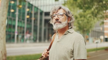 Gri saçlı, sakallı orta yaşlı bir adam yürüyor ve etrafına bakıyor. Omzunda çanta olan gözlüklü olgun bir beyefendi şehir manzarası arka planında meydanda yürüyor.