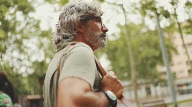 Gri saçlı, sakallı orta yaşlı bir adam yürüyor ve etrafına bakıyor. Omzunda çanta olan gözlüklü olgun bir beyefendi şehir manzarası arka planında meydanda yürüyor.