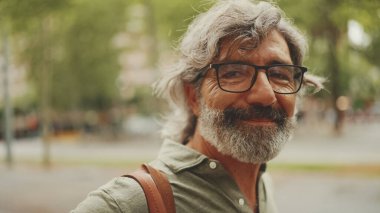 Gri saçlı, sakallı orta yaşlı bir adam yürüyor ve etrafına bakıyor. Omzunda çanta olan gözlüklü olgun bir beyefendi şehir manzarası arka planında meydanda yürüyor.