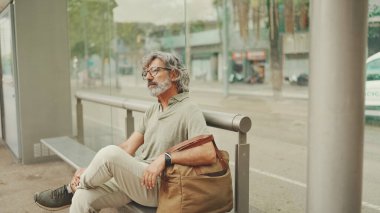 Düşünceli, orta yaşlı, gri saçlı ve sakallı sıradan kıyafetler giyen bir adam, toplu taşıma durağında oturuyor. Gözlüklü ve çantalı bir beyefendi tramvay istasyonunda bekliyor.