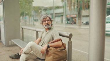 Düşünceli, orta yaşlı, gri saçlı ve sakallı sıradan kıyafetler giyen bir adam, toplu taşıma durağında oturuyor. Gözlüklü ve çantalı bir beyefendi tramvay istasyonunda bekliyor.