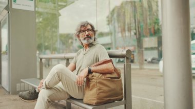 Düşünceli, orta yaşlı, gri saçlı ve sakallı sıradan kıyafetler giyen bir adam, toplu taşıma durağında oturuyor. Gözlüklü ve çantalı bir beyefendi tramvay istasyonunda bekliyor.