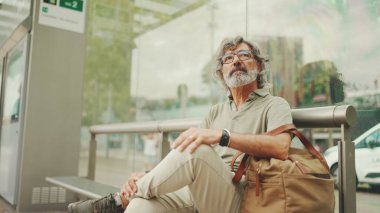 Düşünceli, orta yaşlı, gri saçlı ve sakallı sıradan kıyafetler giyen bir adam, toplu taşıma durağında oturuyor. Gözlüklü ve çantalı bir beyefendi tramvay istasyonunda bekliyor.