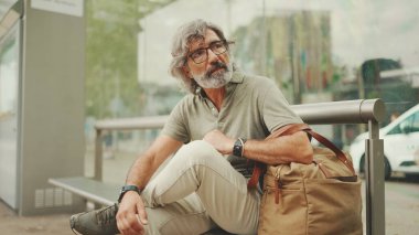 Düşünceli, orta yaşlı, gri saçlı ve sakallı sıradan kıyafetler giyen bir adam, toplu taşıma durağında oturuyor. Gözlüklü ve çantalı bir beyefendi tramvay istasyonunda bekliyor.