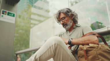 Düşünceli, orta yaşlı, gri saçlı ve sakallı sıradan kıyafetler giyen bir adam, toplu taşıma durağında oturuyor. Gözlüklü ve çantalı bir beyefendi tramvay istasyonunda bekliyor.