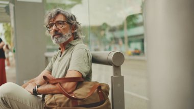 Düşünceli, orta yaşlı, gri saçlı ve sakallı sıradan kıyafetler giyen bir adam, toplu taşıma durağında oturuyor. Gözlüklü ve çantalı bir beyefendi tramvay istasyonunda bekliyor.