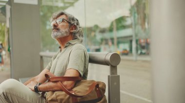 Düşünceli, orta yaşlı, gri saçlı ve sakallı sıradan kıyafetler giyen bir adam, toplu taşıma durağında oturuyor. Gözlüklü ve çantalı bir beyefendi tramvay istasyonunda bekliyor.