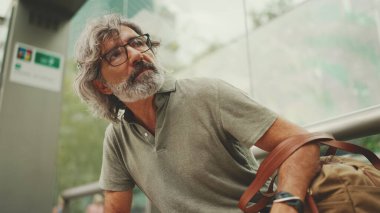 Düşünceli, orta yaşlı, gri saçlı ve sakallı sıradan kıyafetler giyen bir adam, toplu taşıma durağında oturuyor. Gözlüklü ve çantalı bir beyefendi tramvay istasyonunda bekliyor.