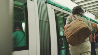 Middle-aged man with gray hair and beard, wearing casual clothes, gets on tram at public transport stop. Mature gentleman in eyeglasses with bag on his shoulder gets on the tram