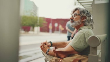 Düşünceli, orta yaşlı, gri saçlı ve sakallı sıradan kıyafetler giyen bir adam, toplu taşıma durağında oturuyor. Gözlüklü ve çantalı bir beyefendi tramvay istasyonunda bekliyor.