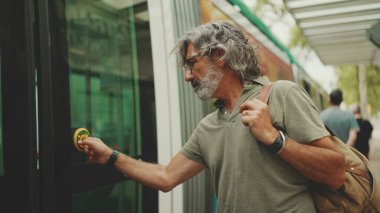 Middle-aged man with gray hair and beard, wearing casual clothes, gets on tram at public transport stop. Mature gentleman in eyeglasses with bag on his shoulder gets on the tram