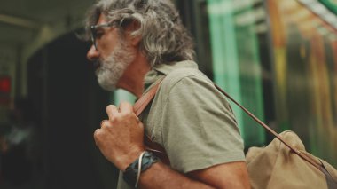 Middle-aged man with gray hair and beard, wearing casual clothes, gets on tram at public transport stop. Mature gentleman in eyeglasses with bag on his shoulder gets on the tram