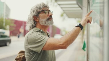 Gri saçlı ve sakallı orta yaşlı adam profili, kentin ulaşım çizelgesinde parmaklarını gezdiriyor. Gözlüklü beyefendi tramvayı bekliyor.