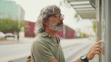 Gri saçlı ve sakallı orta yaşlı adam profili, kentin ulaşım çizelgesinde parmaklarını gezdiriyor. Gözlüklü beyefendi tramvayı bekliyor.