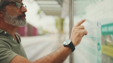 Gri saçlı ve sakallı orta yaşlı adam profili, kentin ulaşım çizelgesinde parmaklarını gezdiriyor. Gözlüklü beyefendi tramvayı bekliyor.