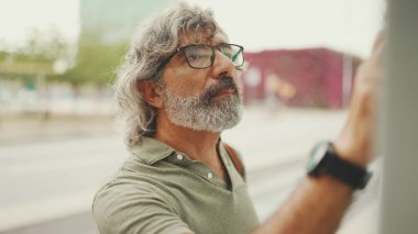 Gri saçlı ve sakallı orta yaşlı adam profili, kentin ulaşım çizelgesinde parmaklarını gezdiriyor. Gözlüklü beyefendi tramvayı bekliyor.