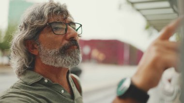 Gri saçlı ve sakallı orta yaşlı adam profili, kentin ulaşım çizelgesinde parmaklarını gezdiriyor. Gözlüklü beyefendi tramvayı bekliyor.