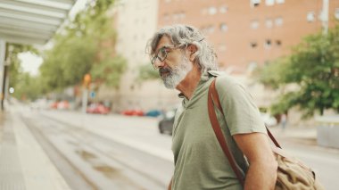 Dost canlısı, orta yaşlı, gri saçlı, sakallı, sıradan kıyafetler giyen bir adam arka planda şehir ulaşımına doğru yürüyor.