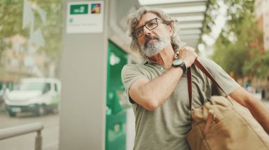Dost canlısı, orta yaşlı, gri saçlı, sakallı, sıradan kıyafetler giyen bir adam arka planda şehir ulaşımına doğru yürüyor.