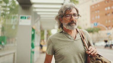 Dost canlısı, orta yaşlı, gri saçlı, sakallı, sıradan kıyafetler giyen bir adam arka planda şehir ulaşımına doğru yürüyor.