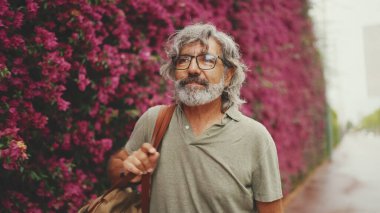 Dost canlısı, orta yaşlı, gri saçlı ve sakallı bir adam, arka planda çiçek açan mor çiçeklerle duvarın yanında yürüyor.
