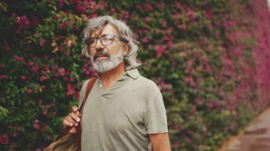 Dost canlısı, orta yaşlı, gri saçlı ve sakallı bir adam, arka planda çiçek açan mor çiçeklerle duvarın yanında yürüyor.