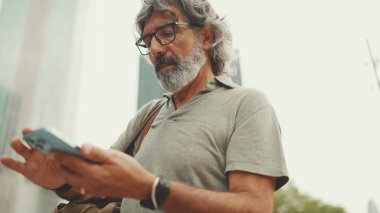 Gri saçlı ve sakallı arkadaş canlısı orta yaşlı bir adam haritaya bakarak cep telefonunu kullanmaya çalışıyor. Gözlüklü, dışarıda cep telefonunda harita uygulaması kullanan olgun bir beyefendi.