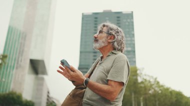 Gri saçlı ve sakallı arkadaş canlısı orta yaşlı bir adam haritaya bakarak cep telefonunu kullanmaya çalışıyor. Gözlüklü, dışarıda cep telefonunda harita uygulaması kullanan olgun bir beyefendi.