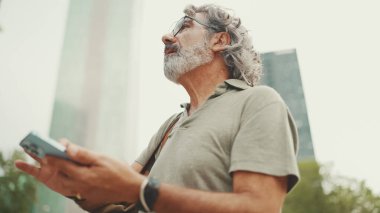 Gri saçlı ve sakallı arkadaş canlısı orta yaşlı bir adam haritaya bakarak cep telefonunu kullanmaya çalışıyor. Gözlüklü, dışarıda cep telefonunda harita uygulaması kullanan olgun bir beyefendi.