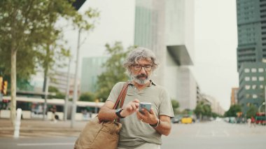 Gri saçlı ve sakallı arkadaş canlısı orta yaşlı bir adam haritaya bakarak cep telefonunu kullanmaya çalışıyor. Gözlüklü, dışarıda cep telefonunda harita uygulaması kullanan olgun bir beyefendi.