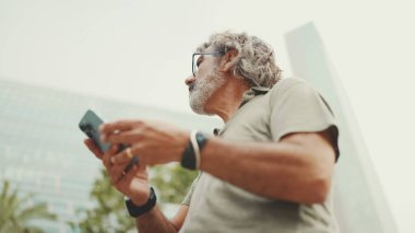 Gri saçlı ve sakallı arkadaş canlısı orta yaşlı bir adam haritaya bakarak cep telefonunu kullanmaya çalışıyor. Gözlüklü, dışarıda cep telefonunda harita uygulaması kullanan olgun bir beyefendi.