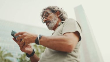 Gri saçlı ve sakallı arkadaş canlısı orta yaşlı bir adam haritaya bakarak cep telefonunu kullanmaya çalışıyor. Gözlüklü, dışarıda cep telefonunda harita uygulaması kullanan olgun bir beyefendi.