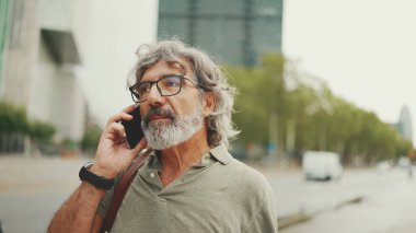 Dost canlısı, orta yaşlı, gri saçlı, sakallı, sıradan kıyafetler giyen ve cep telefonunu kullanan bir adam. Gözlüklü, dışarıda cep telefonuyla konuşan olgun bir beyefendi.