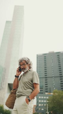 Dost canlısı, orta yaşlı, gri saçlı, sakallı, sıradan kıyafetler giyen ve cep telefonunu kullanan bir adam. Gözlüklü, dışarıda cep telefonuyla konuşan olgun bir beyefendi.