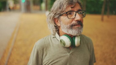 Gri saçlı, sakallı, sıradan kıyafetler giyen, kulaklık takan düşünceli orta yaşlı bir adam sokakta yürüyor.