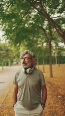 Gri saçlı, sakallı, sıradan kıyafetler giyen, kulaklık takan düşünceli orta yaşlı bir adam sokakta yürüyor.