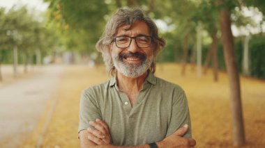 Gri saçlı, sakallı, sıradan kıyafetler giyen, dost canlısı, orta yaşlı bir adam kameraya bakıyor. Gözlüklü olgun beyefendi kameraya gülümsüyor.