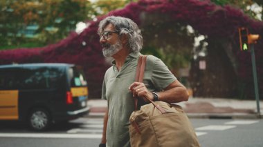 Gri saçlı ve sakallı, samimi, orta yaşlı bir adam yaya geçidinden karşıya geçiyor. Gözlüklü olgun bir beyefendi yaya geçidinden karşıya geçerken arkasına bakıyor.